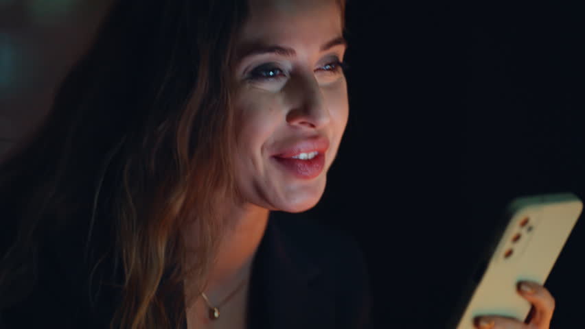 Closeup businesswoman holding smartphone sitting dark office. Happy cheerful woman manager tapping cellphone on evening workplace. Corporate employee texting message mobile phone in modern agency