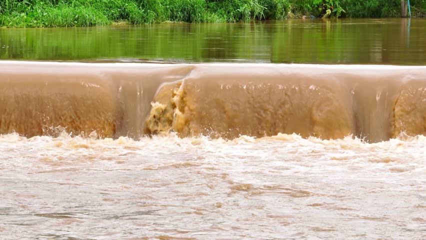 Overhead shot reveals brown floodwaters rushing over a levee, demonstrating the impact of heavy rainfall and flood events on natural water systems and sediment dispersal.
