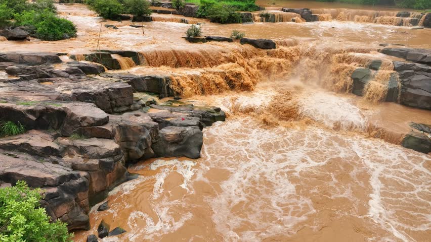 Bird’s eye perspective shows turbulent sediment-rich floodwaters surging through rocks, illustrating the dynamic movement and erosion caused by flooding. Thailand.
