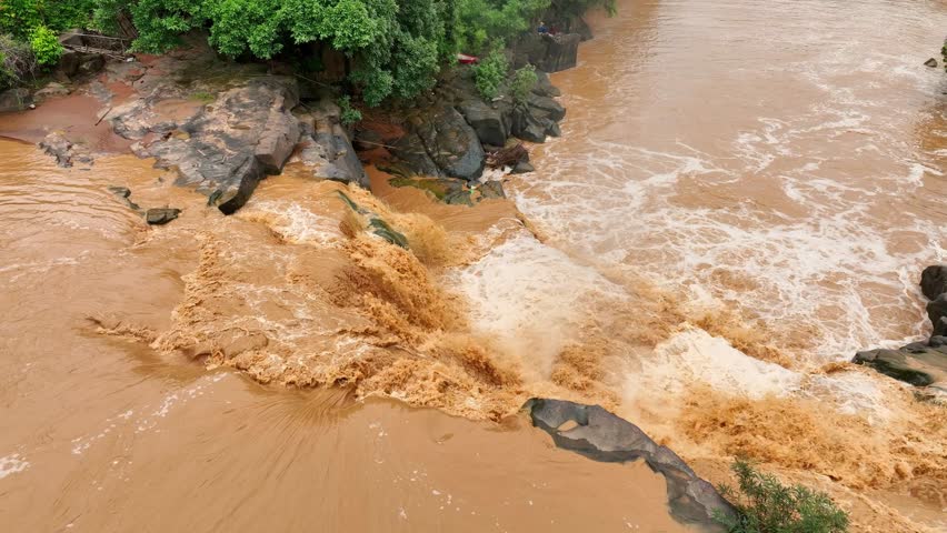 Bird’s eye shot captures turbulent muddy waters cascading through a rocky river, emphasizing floods exacerbated by human activities such as deforestation and poor land use. Thailand.
