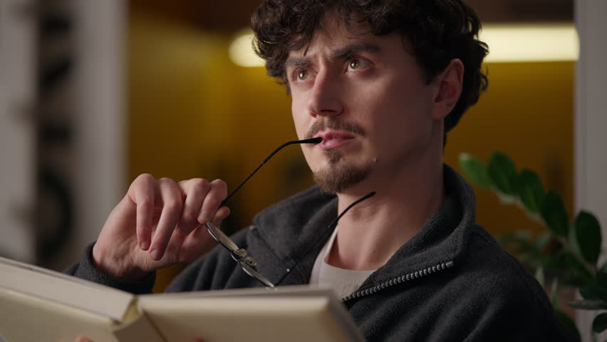 Puzzled pensive Hispanic guy contemplating reading book at home learner read book confused hipster reader learning pages man studying classic literature male lost in thought holding eyeglasses library
