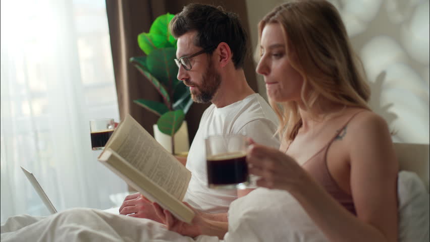 Family calm Caucasian couple drinking coffee in bed bedroom morning relationship wife read book literature pair relaxing bedding man using laptop husband woman drink energizing beverage weekend rest