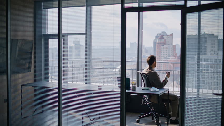 Successful businessman drinking coffee sitting at window luxurious private office. Bearded pensive ceo enjoy hot beverage on work break at workplace. Relaxed man manager in suit looking at cityscape 