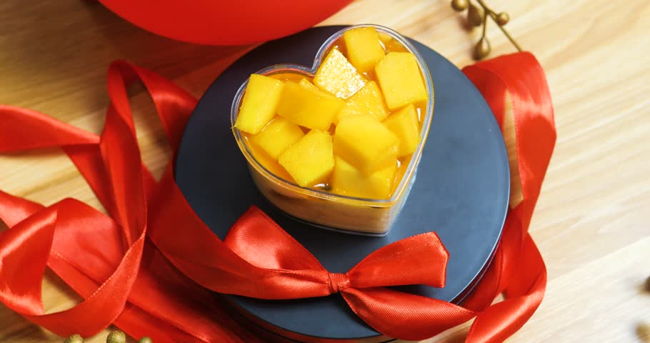 Romantic dessert featuring heart shaped pastry, filled with ripe mango pieces, garnished with red ribbon on dark plate, embodying valentine's day sweetness