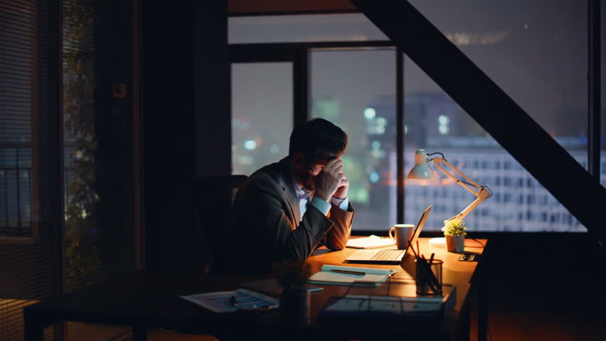Calling executive working night cabinet closeup. Nervous worried man manager talking mobile phone at panoramic dark workplace. Stressed business consultant sitting office desk with laptop writing note