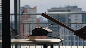 Man hitting old landline phone using wooden bat inside tall glass tower workplace closeup. Pieces explode midair from impact. Skyscraper view appears behind wide transparent window during daytime - Powered by Shutterstock - Get 15% off with code: PIKWIZARD15