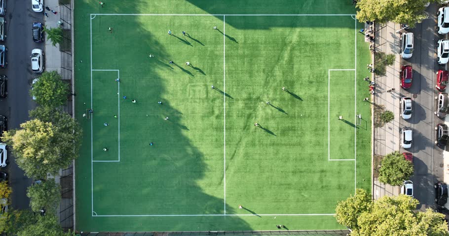 Vertical ascent over small urban soccer field with players scattered across green turf casting long shadows surrounded by street cars buildings trees and community activity in bright afternoon light