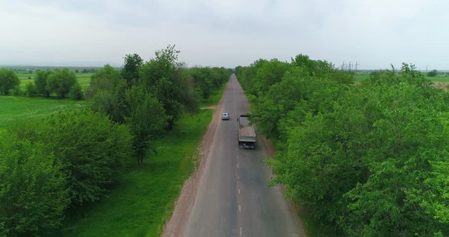 A drone flies over a truck driving on a country road. There are trees and fields around.