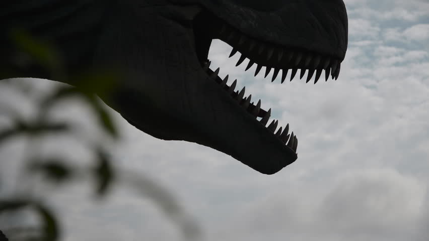 Silhouette of dinosaur head turning head and opening mouth with sharp teeth. Mechanical doll of predatory tyrannosaurus in park against cloudy sky.
