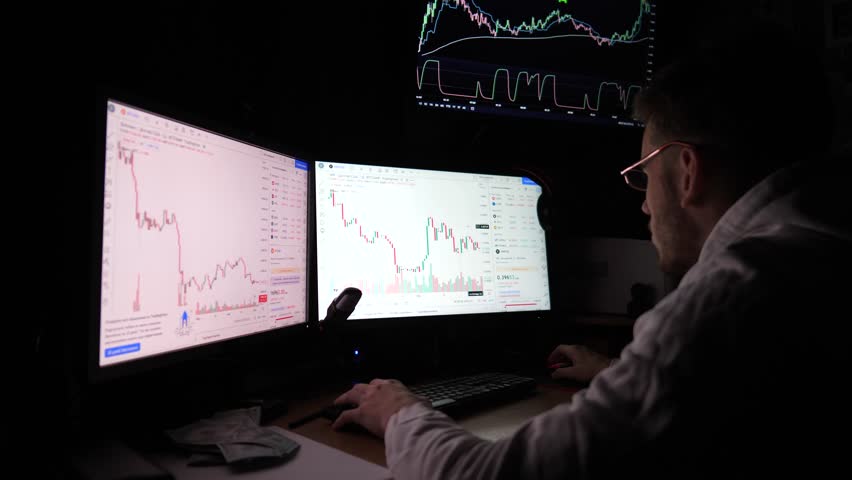 Male trader analyzing commodities and foreign exchange charts. Financial analyst trading online at stock market. Young broker working on computer with multi-monitors at night. Data analysis. Slow mo