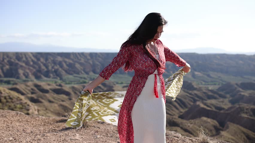 Graceful female dancer wearing red dress swirling silk shawl while moving against dramatic desert canyon landscape during golden sunset light