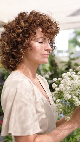 Radiant woman enjoying white flower bouquet, smiling brightly while inhaling fragrant blossoms at sunny farmers market. Vertical