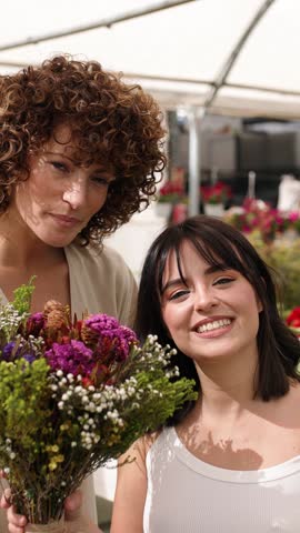 Two cheerful women holding and smelling a colorful bouquet of flowers at a flower shop or market. Vertical