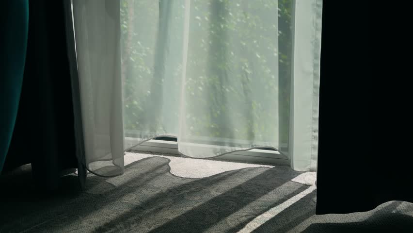 Wind blowing white curtain from open window in hotel. Breeze drifting through open glass and moving sheer drape in sunny interior. Air flowing in from outside and swaying light curtain over soft floor