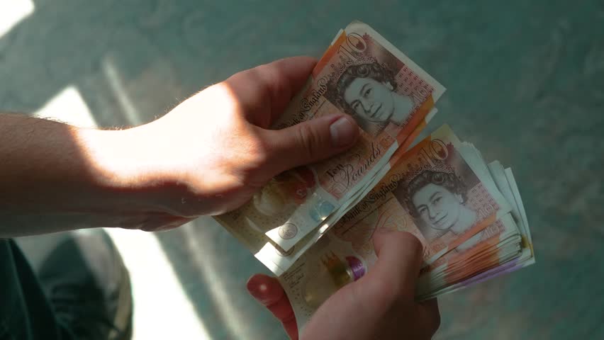 Counting ten pound UK banknotes in daylight. Flipping British currency notes under sunlight. Handling paper money indoors near window. Managing stack of pounds with focused hands