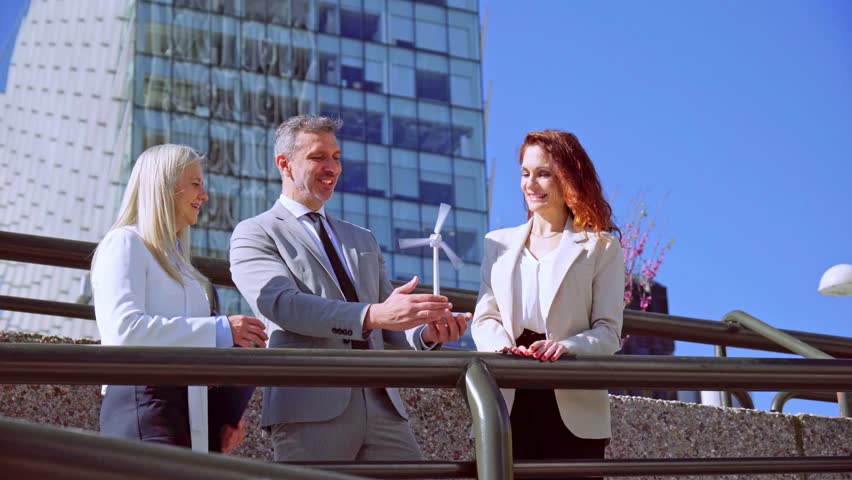 Business professionals examining miniature wind turbine, representing sustainable investment near contemporary skyscraper backdrop with clean energy planning