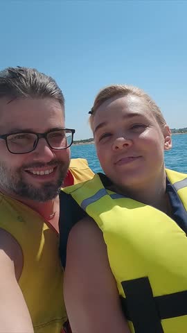 Happy family look at camera and laugh while riding banana boat on blue sea on summer day. Water entertainment attraction. Travel holiday. Vertical shot