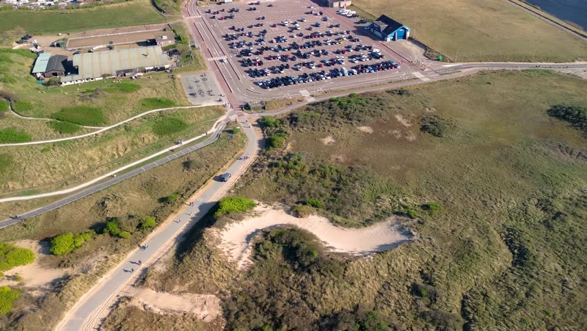 Aerial view of a large parking lot near a river, surrounded by green fields, a skate park, horse stables, and a camping site, with a cityscape in the background under clear skies.