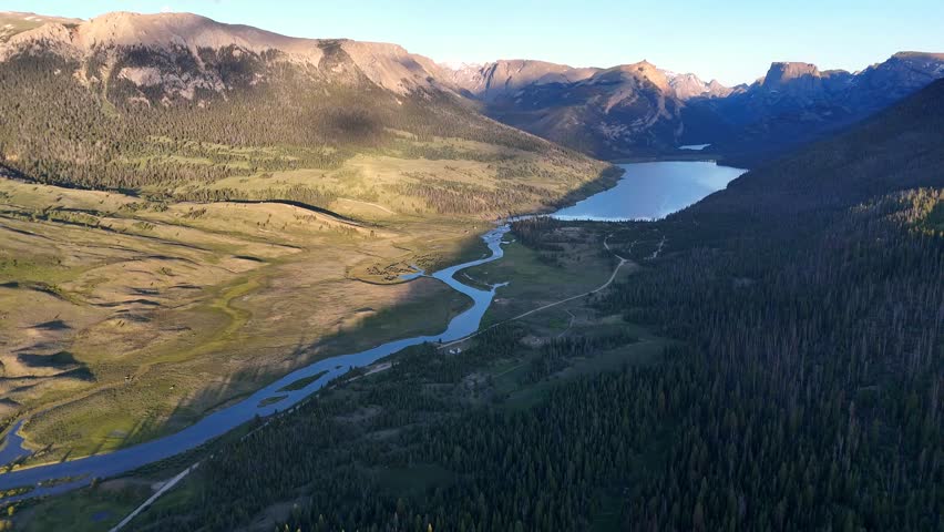 Aerial drone shows winding river flowing through green valley into large lake between tall forest and rocky mountain in scenic peaceful West Virginia landscape under clear blue sky in daylight