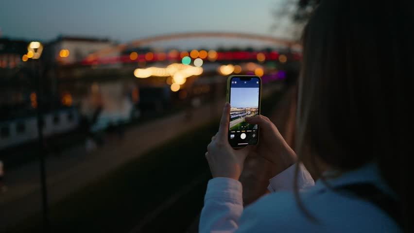 Travel blogger with smartphone captures moments on bridge at night, moments of city lights sparkling, moments of urban beauty. Blogger uses smartphone to save urban memories. Ideal for nostalgic clips