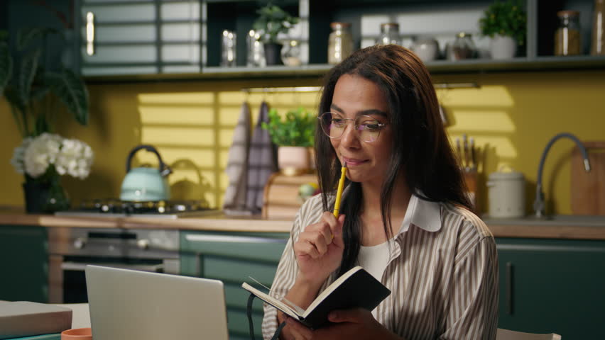 Arabian Indian woman girl businesswoman student freelancer learning studying online class teaching writing notes exam distant education laptop video call chat computer lecture notebook at home kitchen