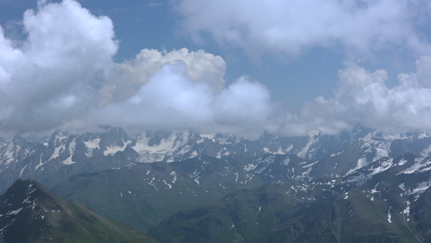 Camera Pans From Left To Right Across A Majestic Mountain Range With Snow-Capped Peaks And Glaciers Under A Blue Sky With Clouds. Scenic Alpine Landscape.