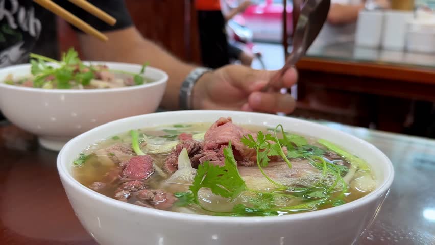 Close-up of steaming bowls of Pho, a traditional Vietnamese beef noodle soup. Fresh herbs and tender beef are visible in the rich broth. Delicious and aromatic!