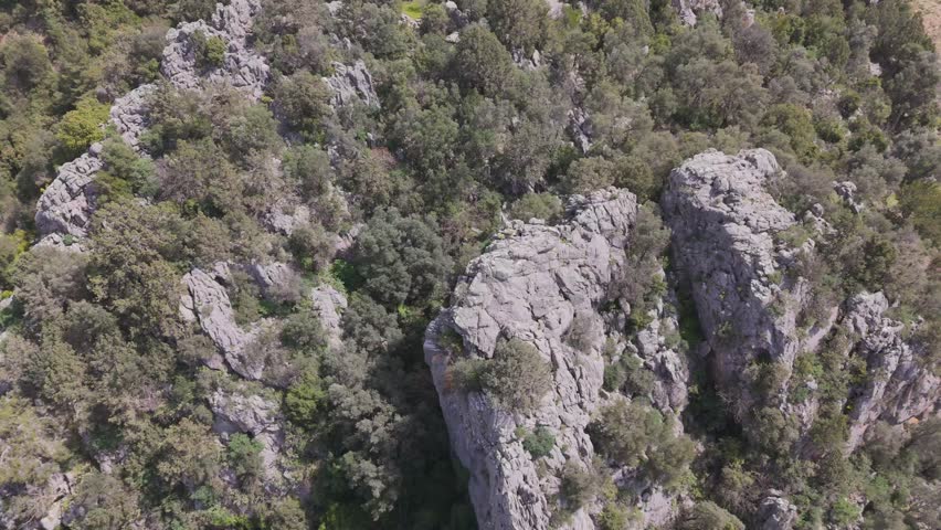 A closeup drone view of forested mountain with rocks and outcrops, overlooking some cottages and winding trail on a sunny day in Hasdumen, Antalya