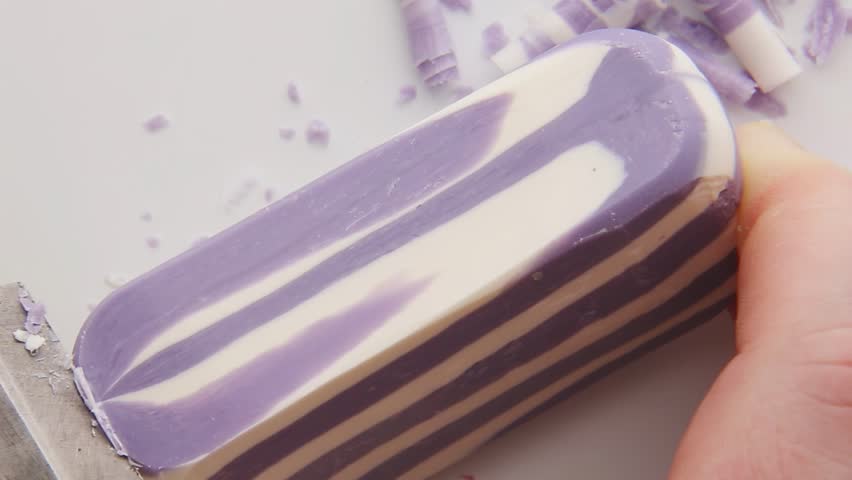 Purple striped soap creates beautiful curling strips as knife peels thin layers producing mesmerizing spiral patterns and therapeutic ASMR sounds on wooden surface creating satisfying experience