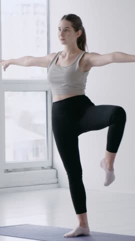 Active sporty brunette girl in sportswear performs a warm-up exercise in a fitness studio. Woman Keeping Body Balance On One Leg While Rotating The Other. Concept of healthy lifestyle. Slow motion