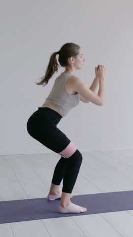 Healthy slim caucasian girl in black leggings and light grey top doing squats with a fitness elastic band on her legs. Concept of sporty female in a studio, Limbering-Up. Slow-mo shot
