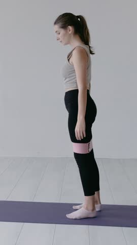 Motivated sporty attractive woman in sportswear doing squats with one leg forward and the other back with a fitness elastic band on legs in a bright sport studio. Young girl stretching legs indoors.