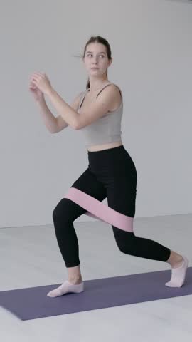 Attractive happy caucasian woman in sportswear doing squats with a fitness elastic band on her legs. Concept of sporty girl in a studio, warming-up exercises. Young girl stretching legs indoors.