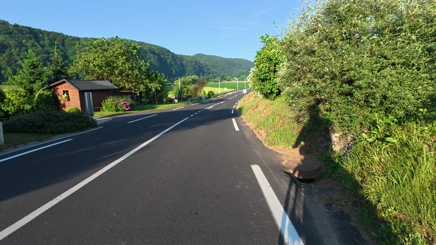 Cyclist riding downhill on a highway in Slovenia, surrounded by scenic mountain landscapes. Shadows falling on the road, creating dynamic light contrasts. 