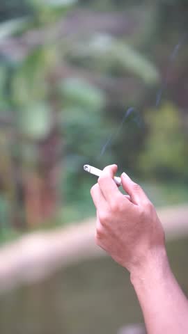 a man's hand holding a burning and smoking clove cigarette, outdoors in the open	