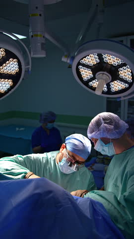 Male surgeons operate under two round lamps. Bearded doctor uses metal tools at work. Vertical video.