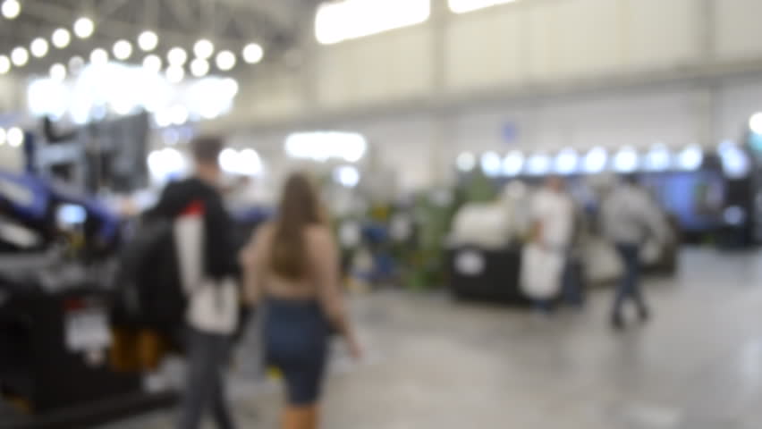 People walking in building. Building with walls, floor, ceiling illuminated lamps. Silhouettes of people indoors. Many people walking. Men and women in tall hall. Exhibition center. Blurred background