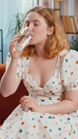 Thirsty woman holding glass of natural aqua make sips drinking still water preventing dehydration sits at home living room. Girl with good life habits, healthy slimming, weight loss concept. Vertical