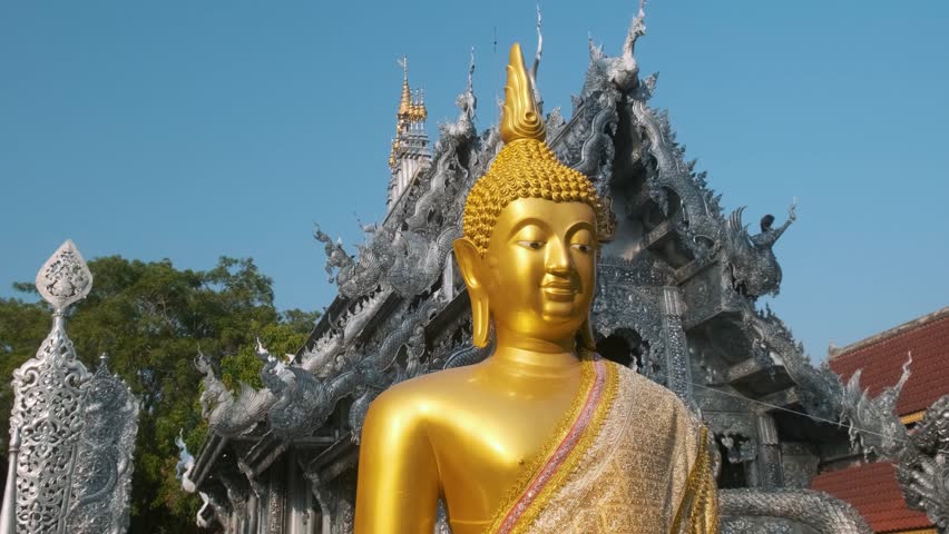 Golden Buddha statue in front of ornate Silver Temple with intricate dragon carvings and detailed metalwork under clear blue sky in Chiang Mai, Thailand. Wat Sri Suphan. Asian architecture