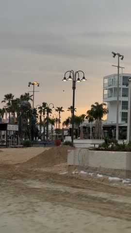 Evening view of a Cyprus beach promenade with public art sculptures, palm trees, modern buildings, and glowing street lamps at sunset.
