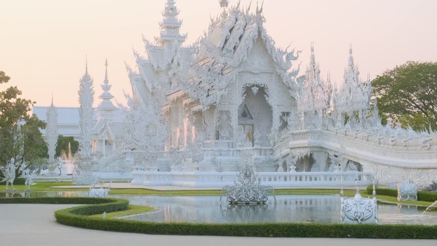 White temple Wat Rong Khun surrounded by lush garden and pond at sunset, Chiang Rai, Thailand. Buddhist temple in Northern Thailand. Travel and touristic landmark in Asia