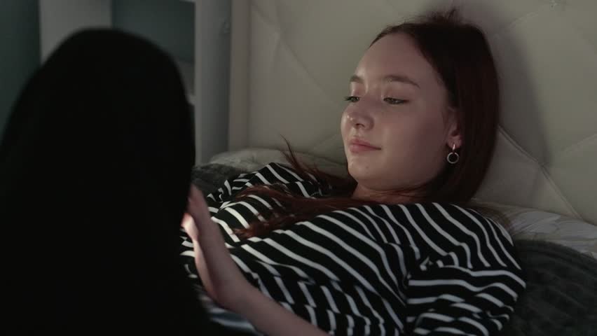 Teenage girl lounging on bed at night, softly illuminated, casually scrolling through smartphone, immersed in digital entertainment and relaxation