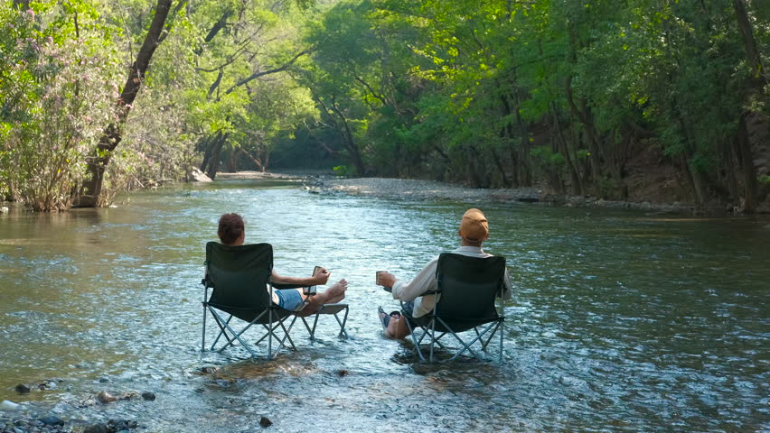 Tourists relaxing in river with feet in water, enjoying drinks. Tourists relaxing on camping chairs, immersed in river waters, savoring refreshing drinks amidst lush forest scenery during vacation