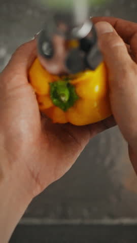 Natural male hands are washing a bright yellow bell pepper under running water in a modern kitchen. This action promotes cleanliness and preparation for healthy cooking