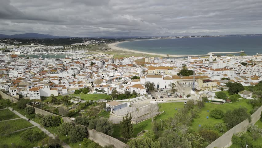Drone flies to the left from southwest corner of old city wall to the northwest corner, looking over the old city on cloudy day in Lagos, Portugal