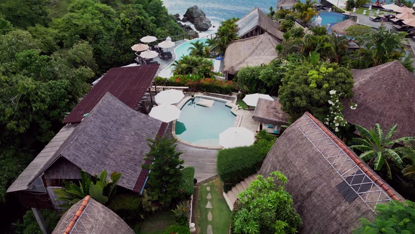 Aerial drone footage captures a stunning tropical resort in Bali perched on a cliffside surrounded by lush nature and endless ocean views offering a peaceful and luxurious island retreat experience.