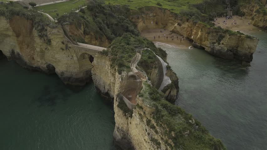 Drone orbits high over stone bridge connecting two rock formations next to beaches and along coast in Lagos, Portugal
