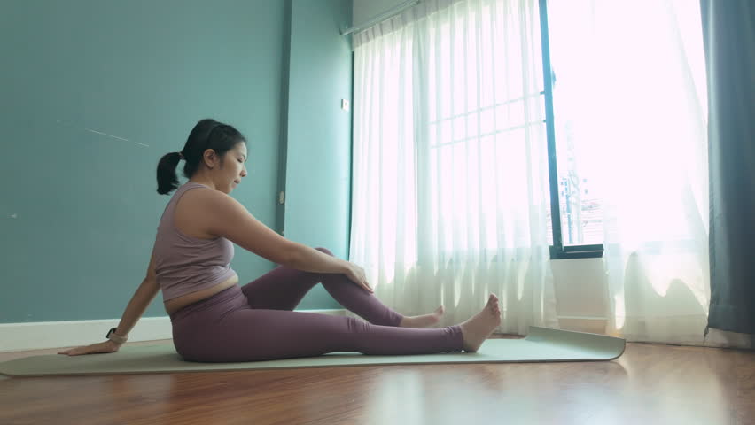 Asian woman performing a Seated Spinal Twist yoga pose on a mat in a bright room, showcasing a peaceful atmosphere.	