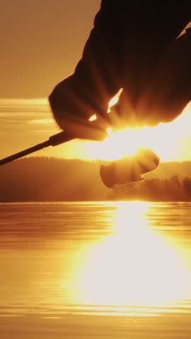 Close-up of a rotating fishing reel in the rays of the sun. A fisherman is fishing by spinning on a lake at sunset. Sunlight breaks through the rotating coil. Vertical video, shorts.
