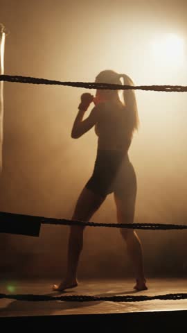 Sport and fitness, beautiful woman punching punch bag in boxing ring, vertical video. Lady training alone in dark gym in night, over trained female person, keep fit and stay slender in middle age